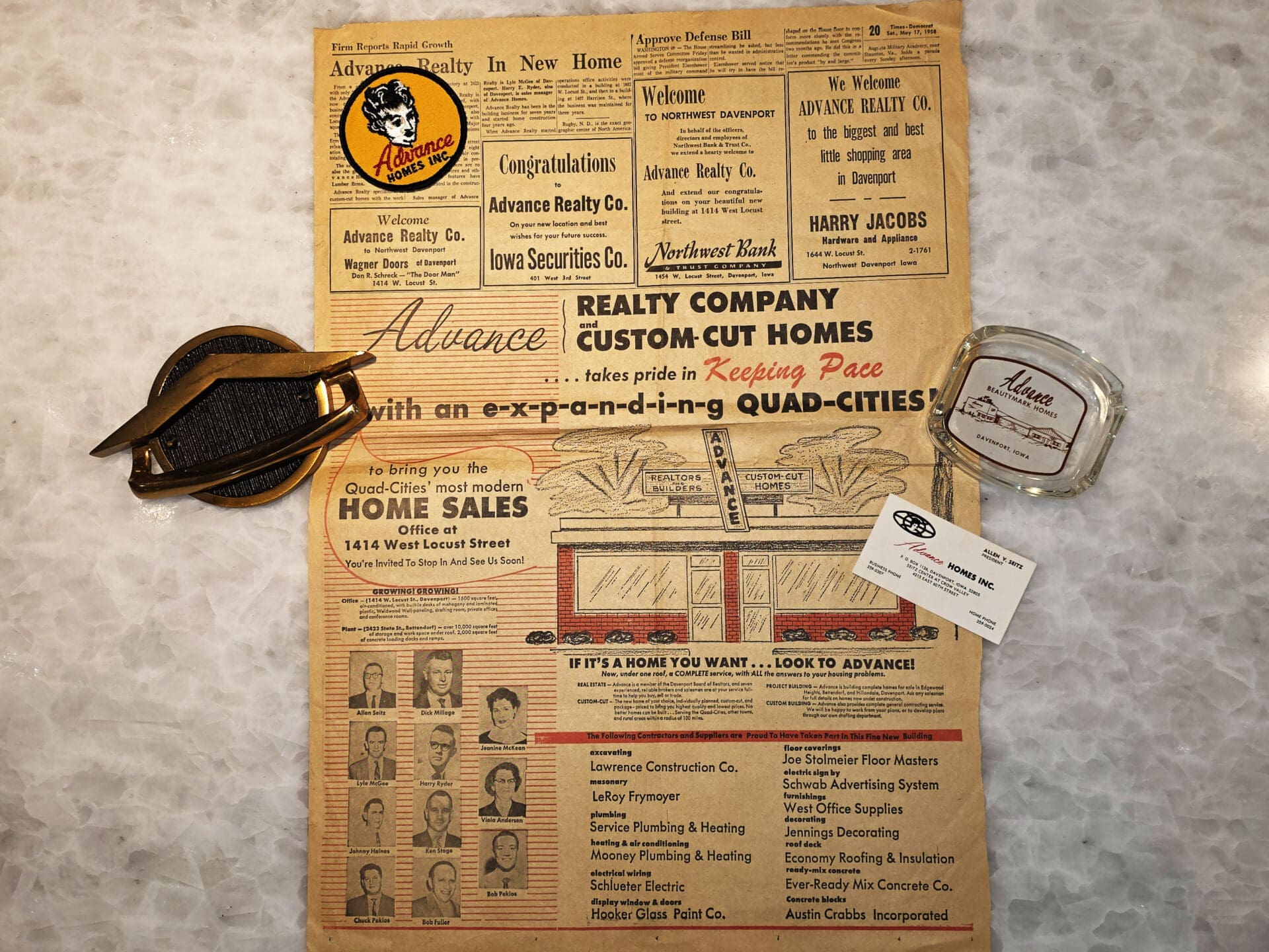 Vintage Advance Homes newspaper ad laid flat on a marble surface, showing headline “Realty Company and Custom-Cut Homes,” illustrated model home, staff portrait boxes, contact details for home sales in the Quad Cities, plus period promotional items and a business card promoting Advance Realty/Advance Homes.