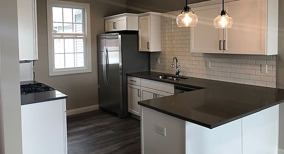 Modern single-family kitchen in a newly built home by Advance Homes: white shaker cabinets, dark quartz countertops with a breakfast bar, subway tile backsplash, stainless steel refrigerator and sink, pendant lights, and wood-look flooring.